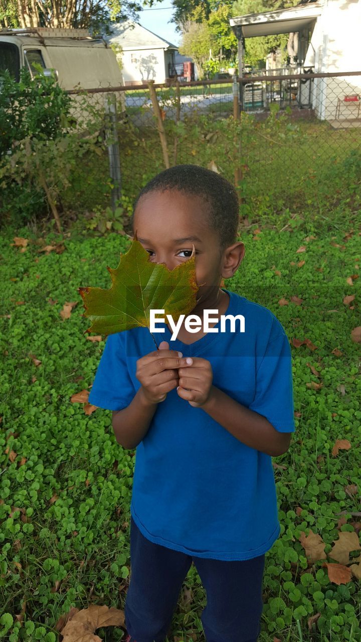 Portrait of boy hiding behind leaf on grassy field