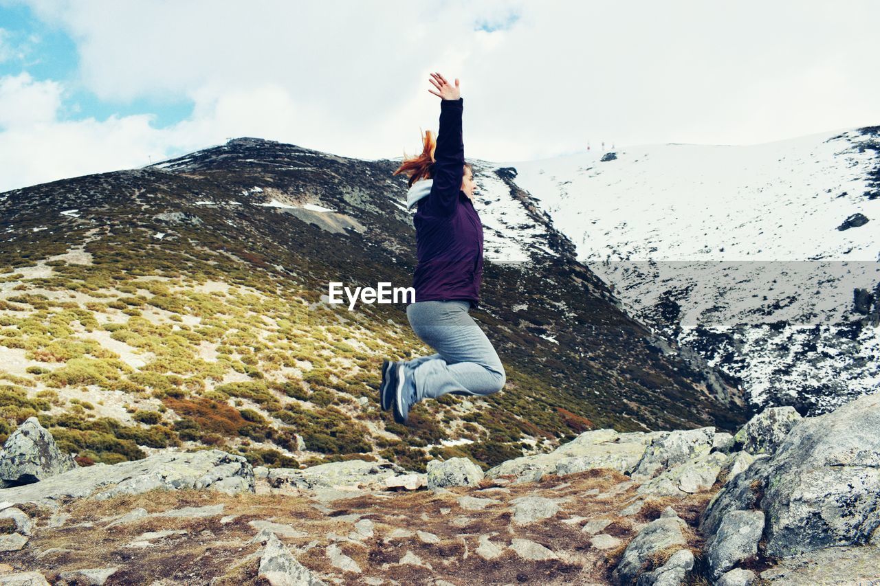 Side view of woman jumping with arms raised on mountain during winter
