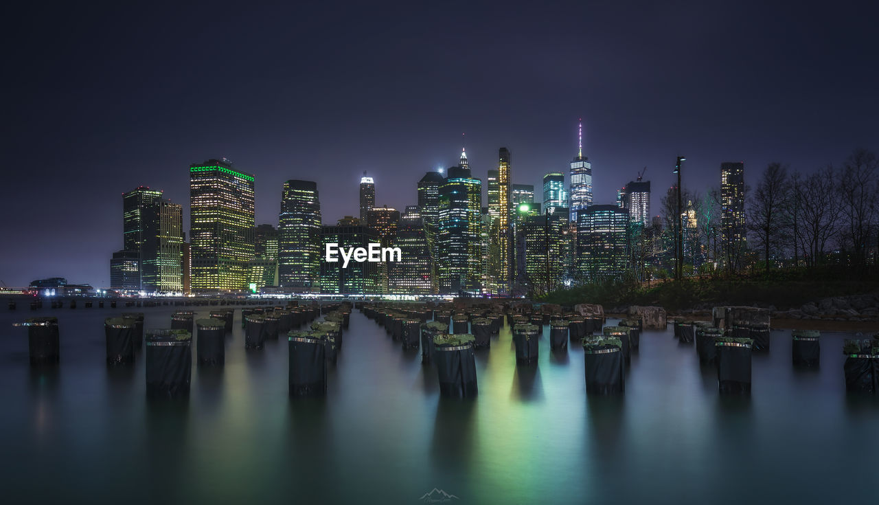 Illuminated modern buildings by river against sky at night