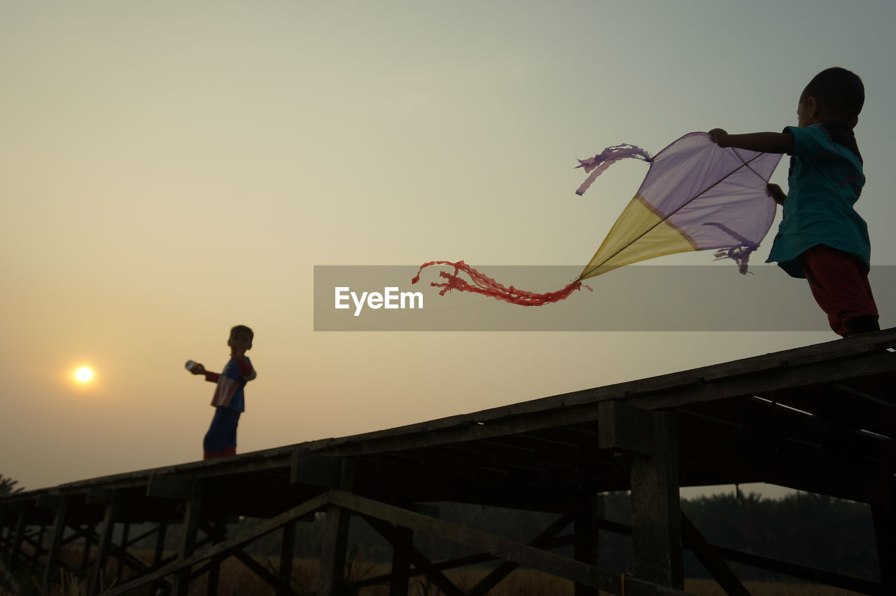 sky, men, sunset, nature, sports, childhood, kite - toy, child, leisure activity, adult, full length, silhouette, extreme sports, low angle view, two people, motion, flying, mid-air, lifestyles, architecture, women, outdoors, jumping, person, copy space, clear sky, standing, activity