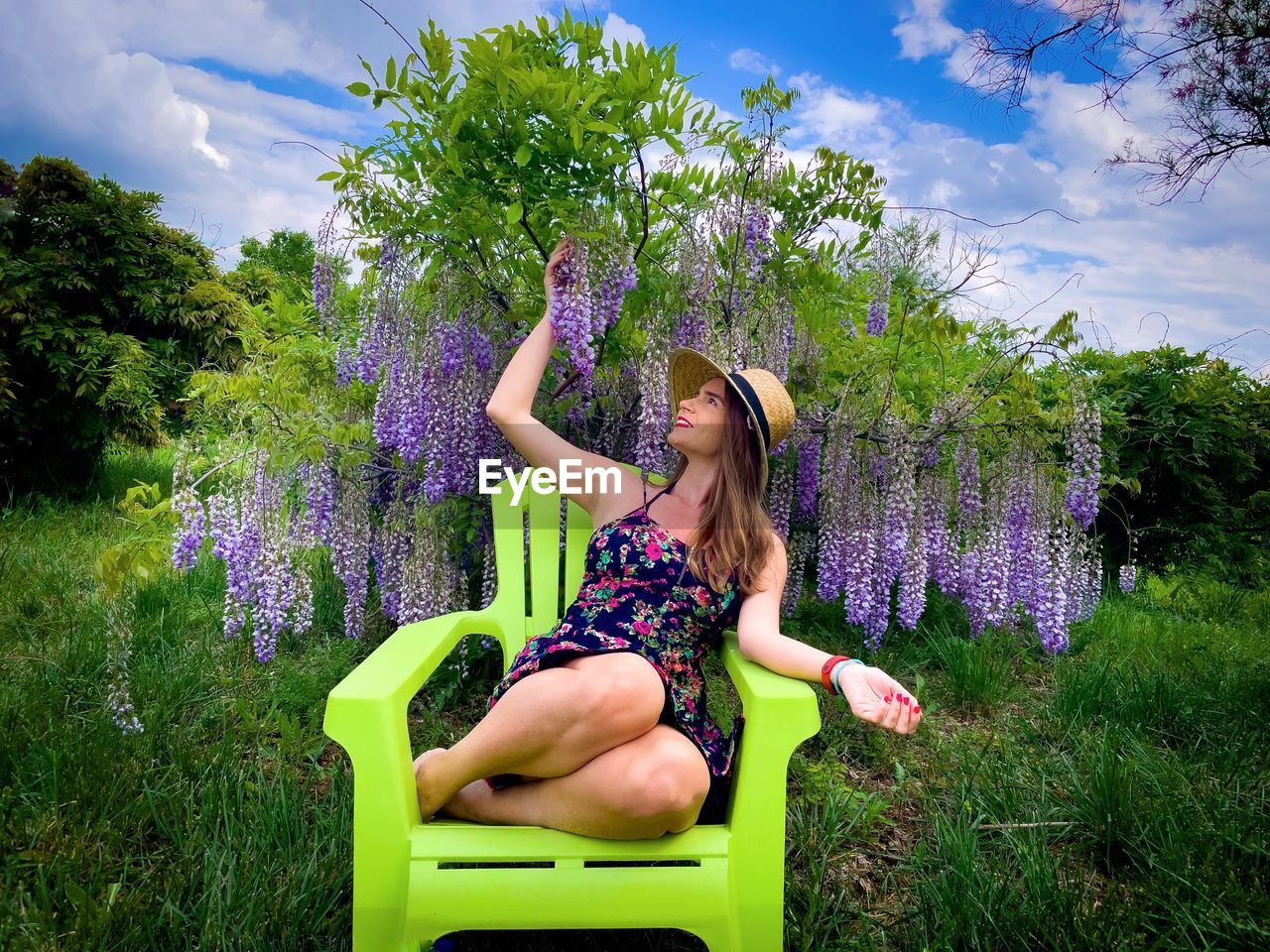 Young woman wearing dress and hat sitting down on a green chair near wisteria flowers