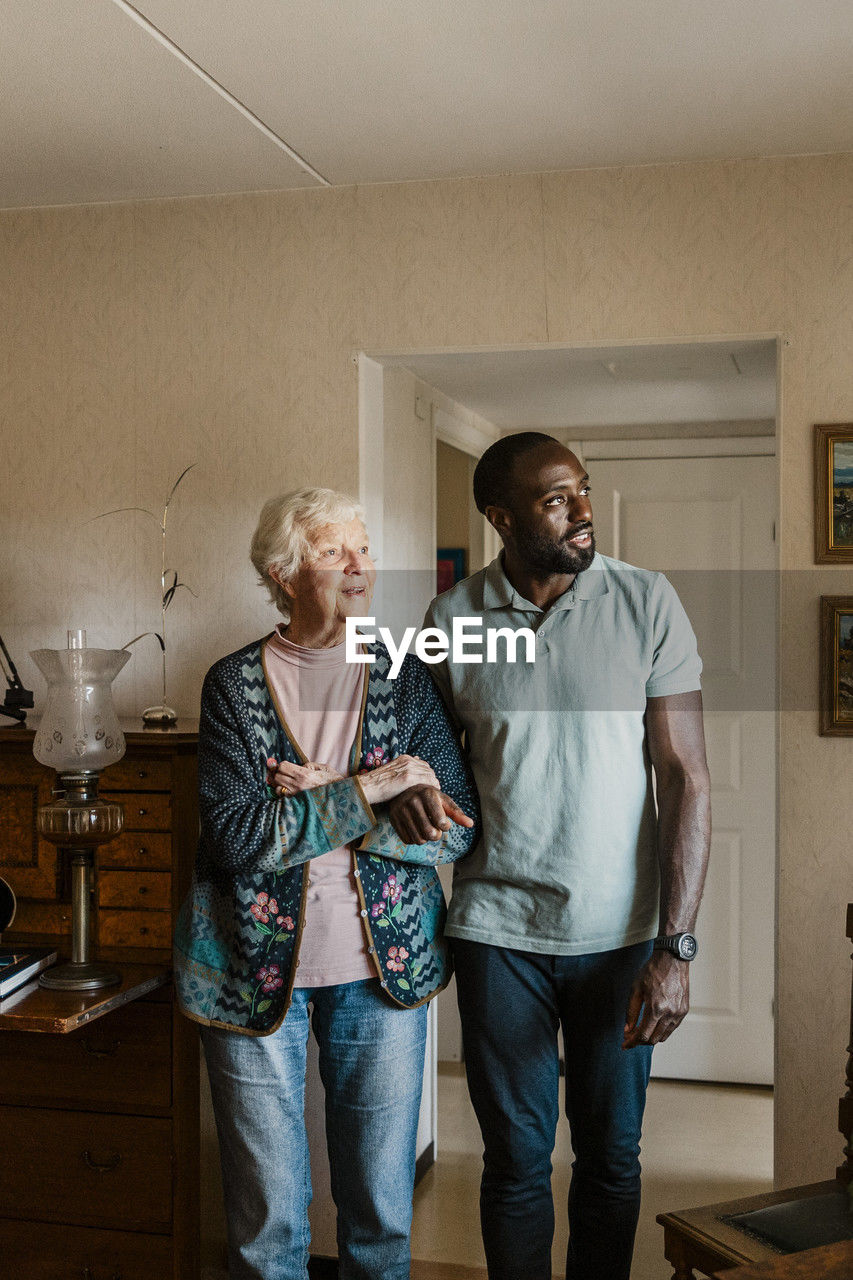 Happy male caretaker walking arm in arm with senior woman in living room at home