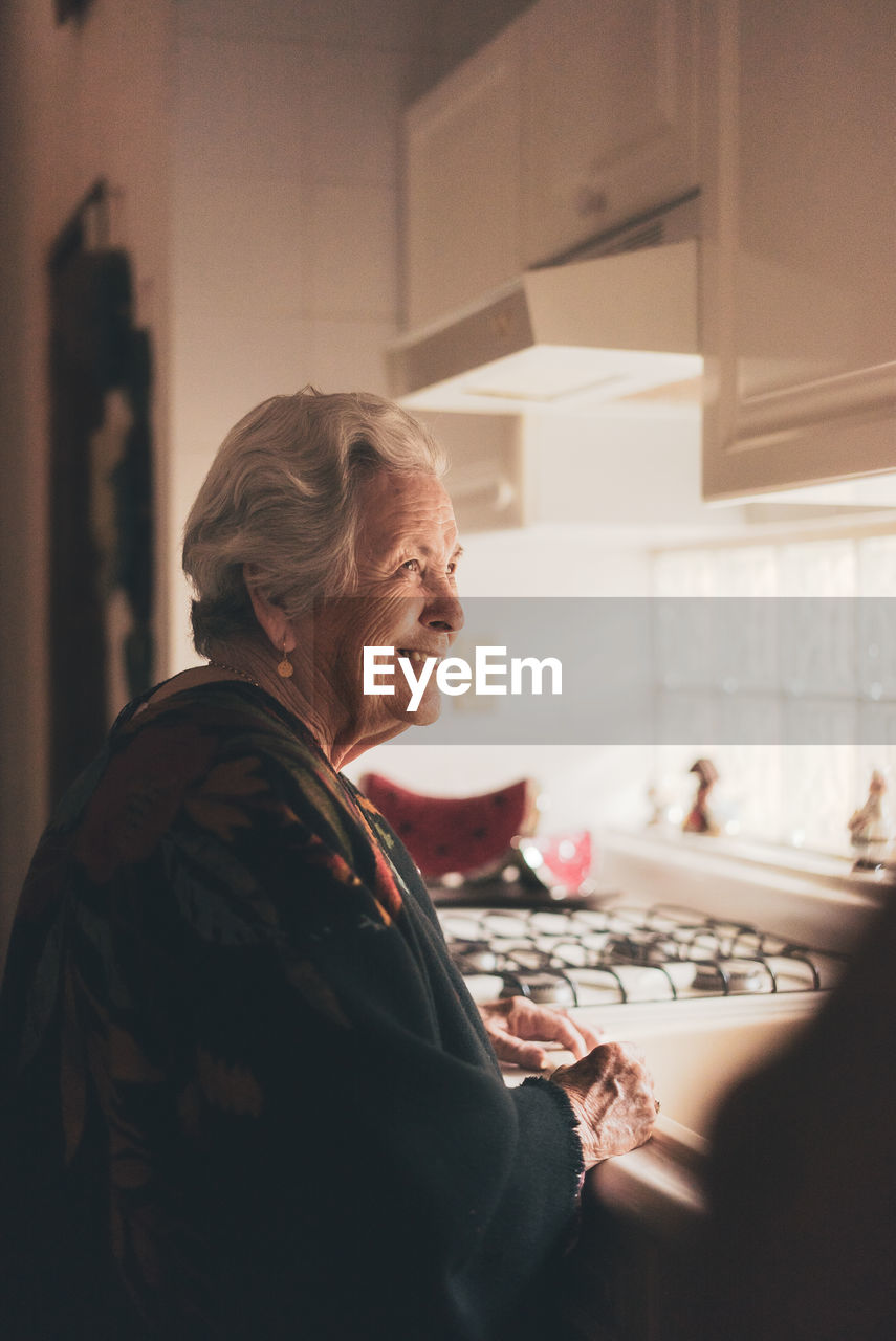 Side view of happy positive senior female with gray hair wearing warm clothes standing at sink in kitchen