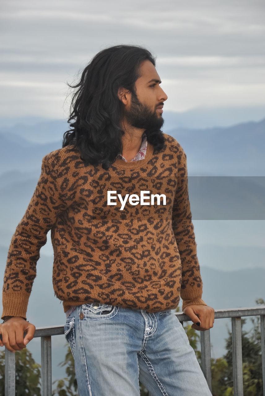 A long haired and bearded indian young man with looking sideways while standing in hilly area 