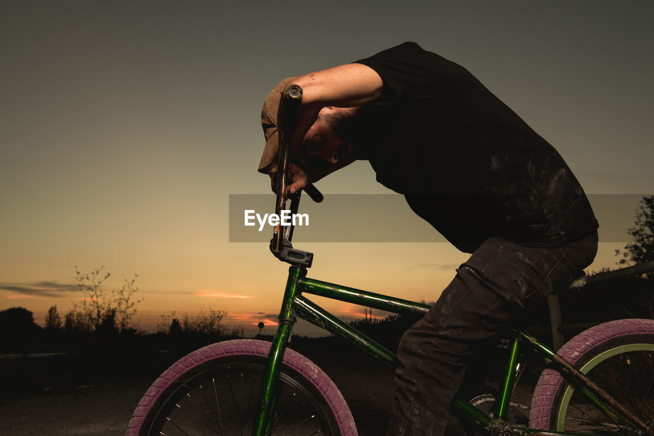 Side view of young man with bicycle sitting on road against sky during sunset