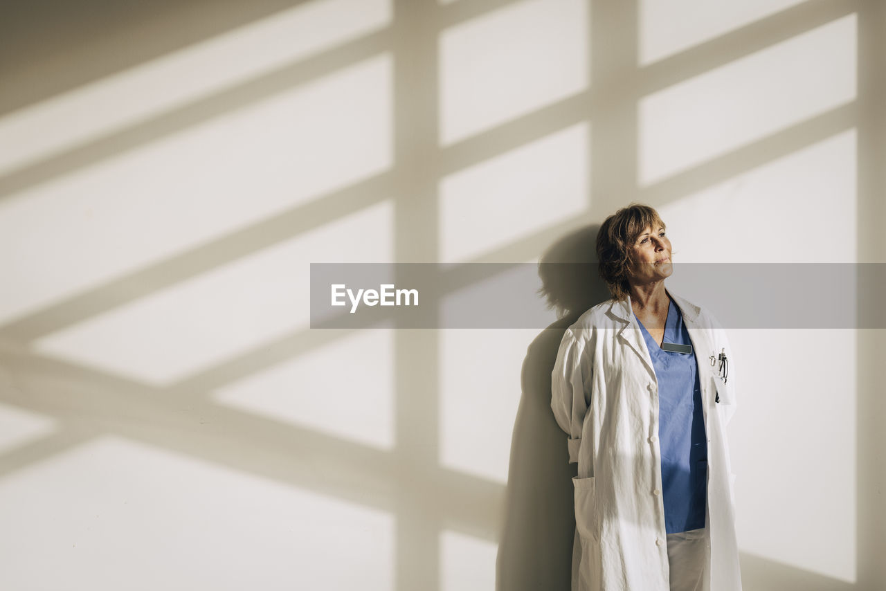 Female doctor wearing lab coat day dreaming while standing against wall with sunlight