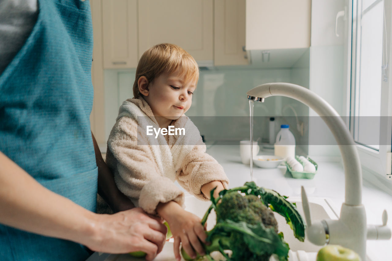 Little cute toddler daughter helps mom to wash vegetables in the kitchen in the sink.