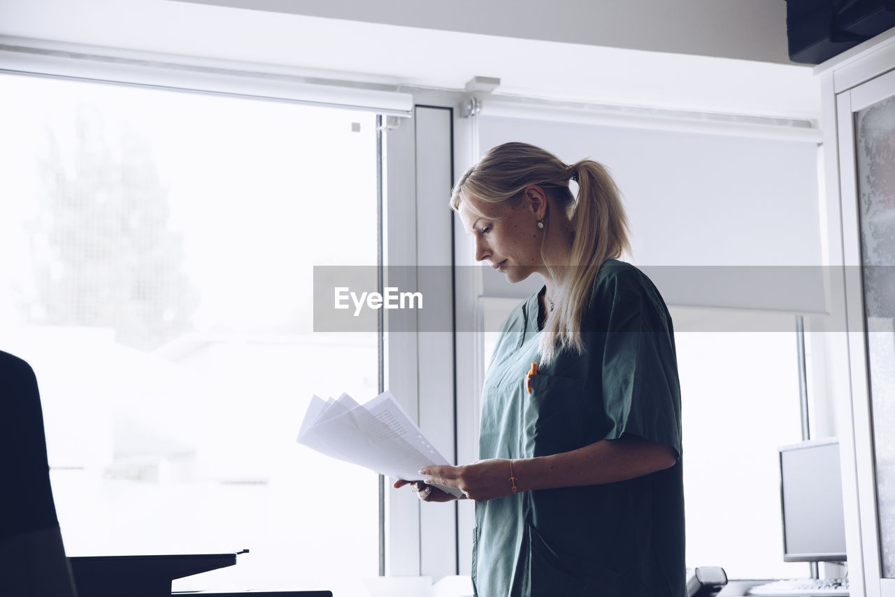 Female nurse reading documents while standing against window at doctor's office