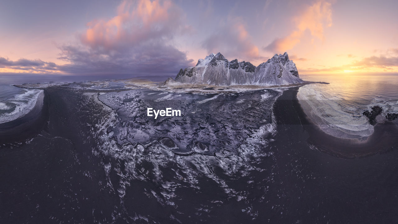 Drone view of calm frozen endless sea surrounding high rocky snowy vestrahorn mountain during picturesque sunset in stockness beach, iceland