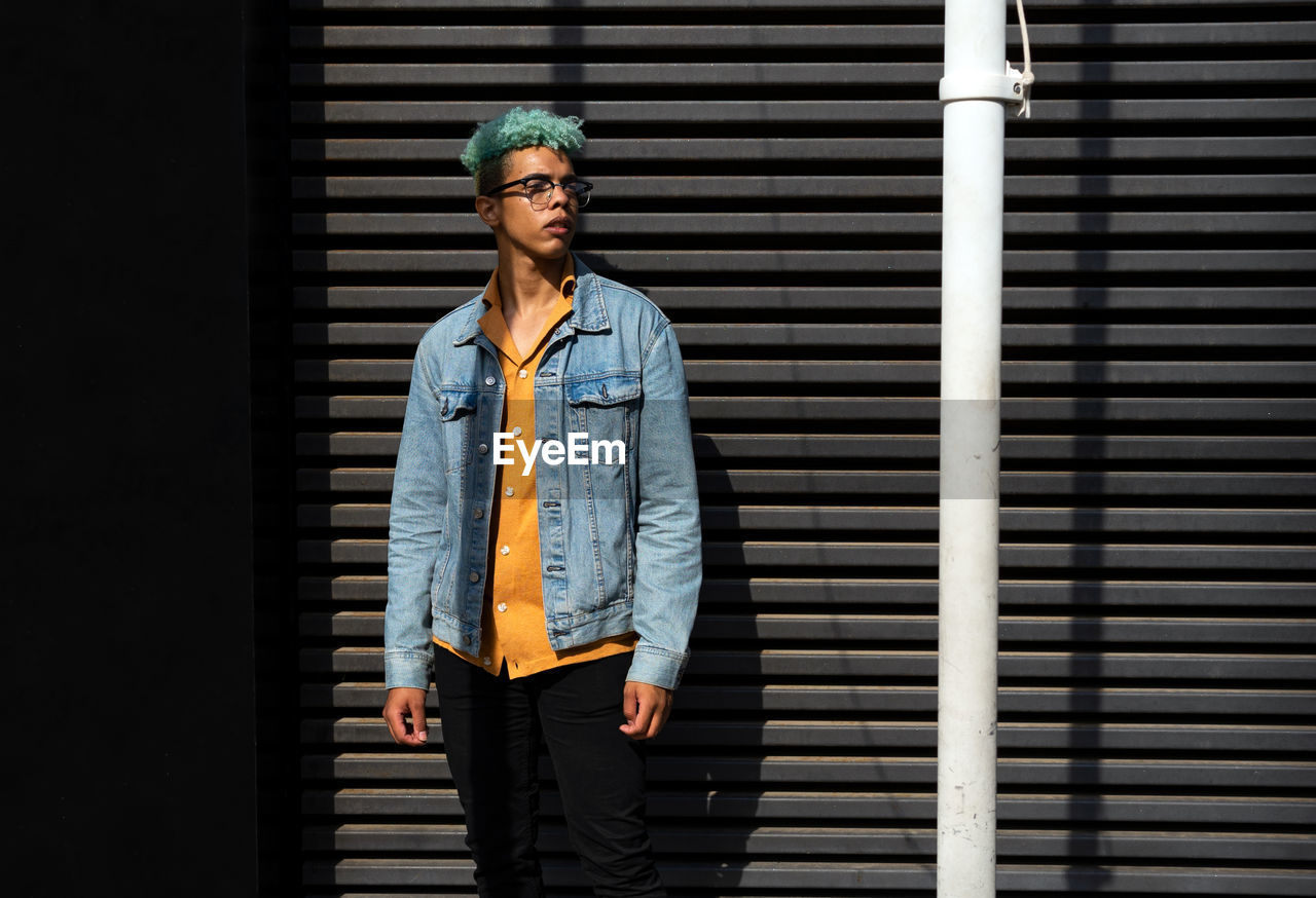 Dreamy black male hipster with blue hair and eyeglasses standing on street and enjoying sunny day looking away