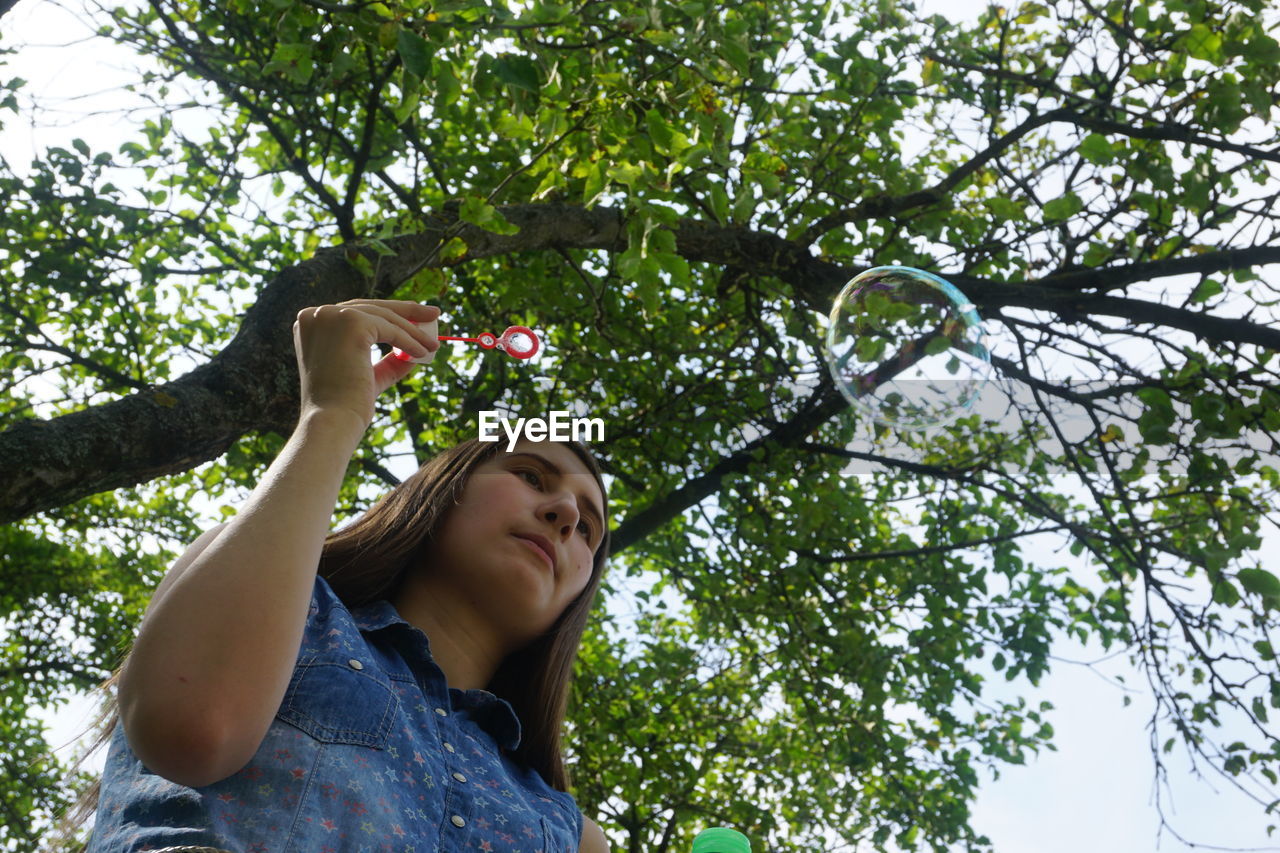 Low angle view of young woman blowing bubbles against tree
