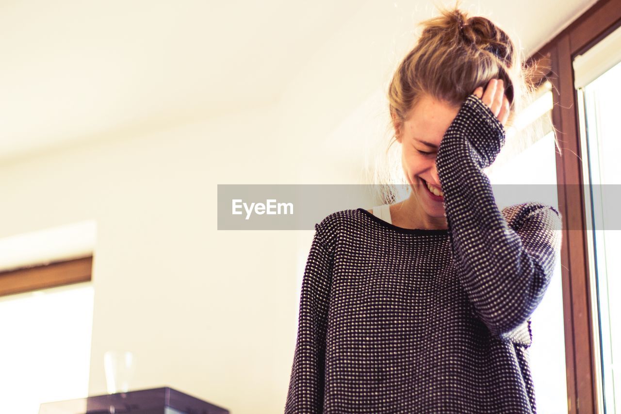 Smiling young woman rubbing eye while standing against wall