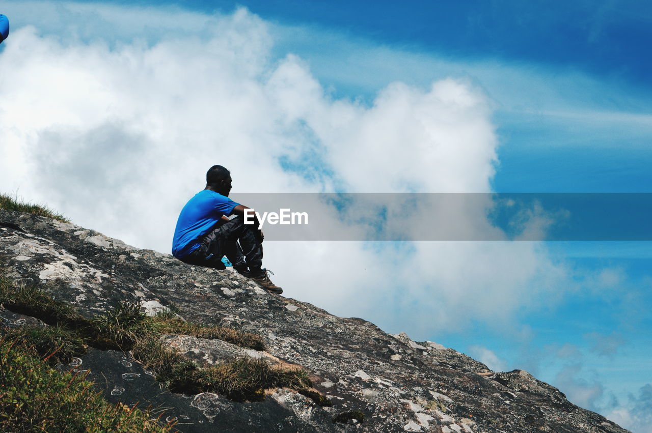 Low angle view of man sitting on cliff against blue sky