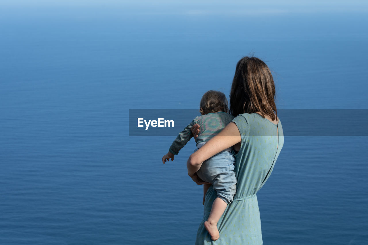 Rear view of woman with daughter standing against blue sky