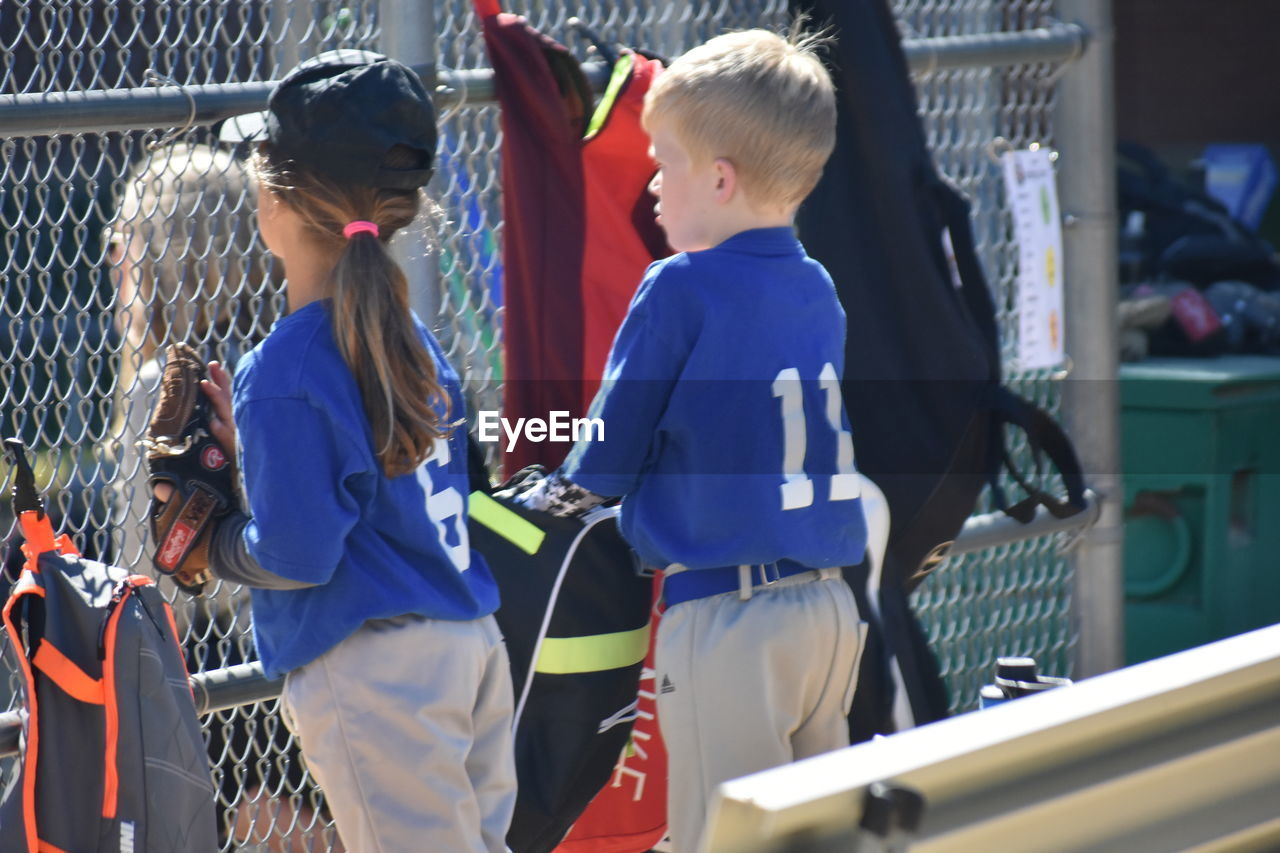 Boy and girl in sports jersey standing by chainlink fence