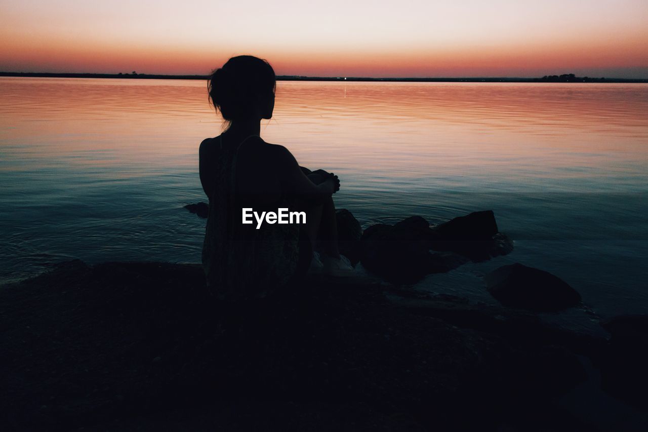 Rear view of woman sitting on rock at seashore during sunset