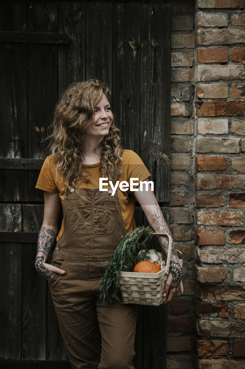 Smiling woman holding vegetable basket against wall