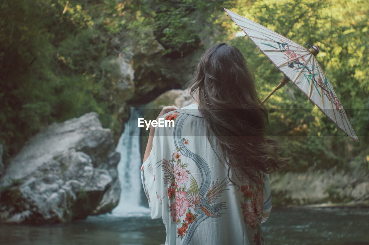 Rear view of woman with umbrella standing by river in forest