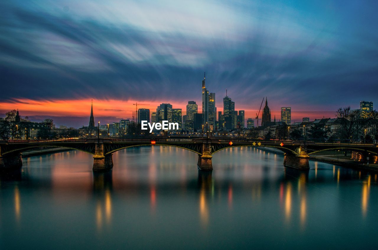 Bridge over river by illuminated buildings against cloudy sky at sunset