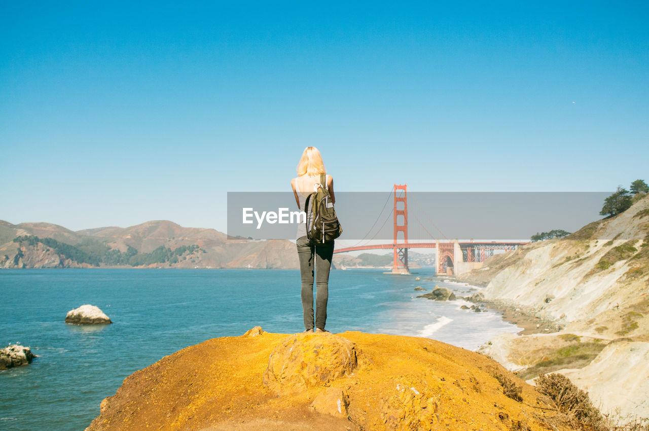 Full length rear view of woman standing on rock at sea shore by golden gate bridge against clear blue sky