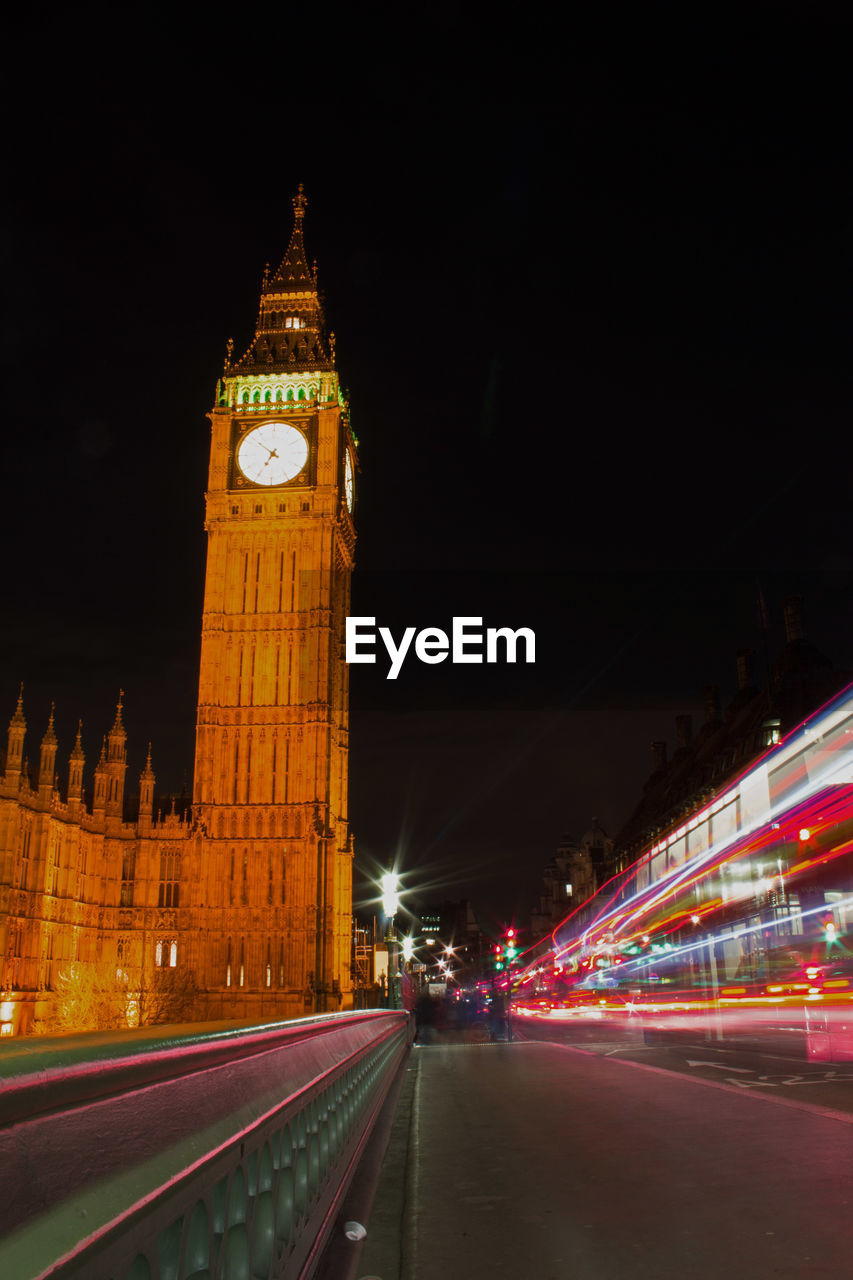 Light trails over street by big ben against sky