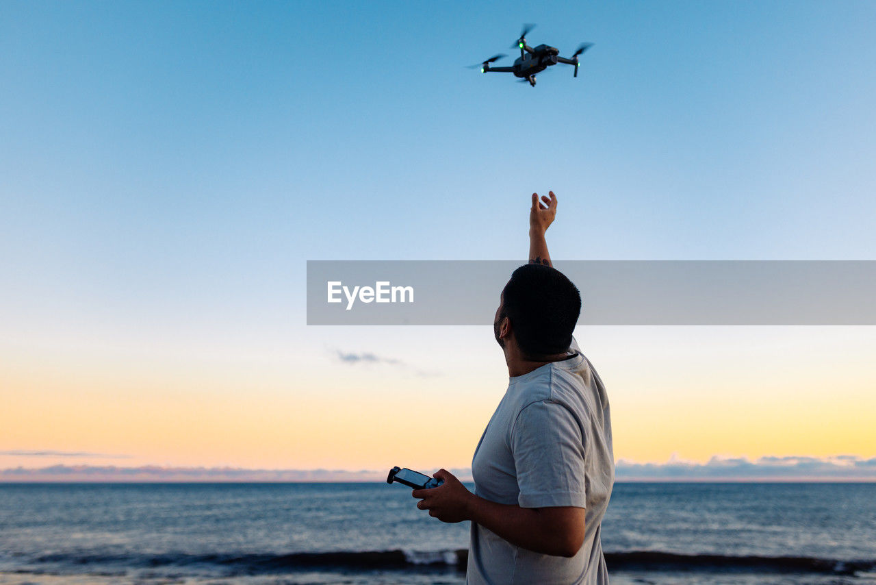 Unrecognizable black male in casual clothes launching modern drone against sunset sky while spending time on beach near waving sea