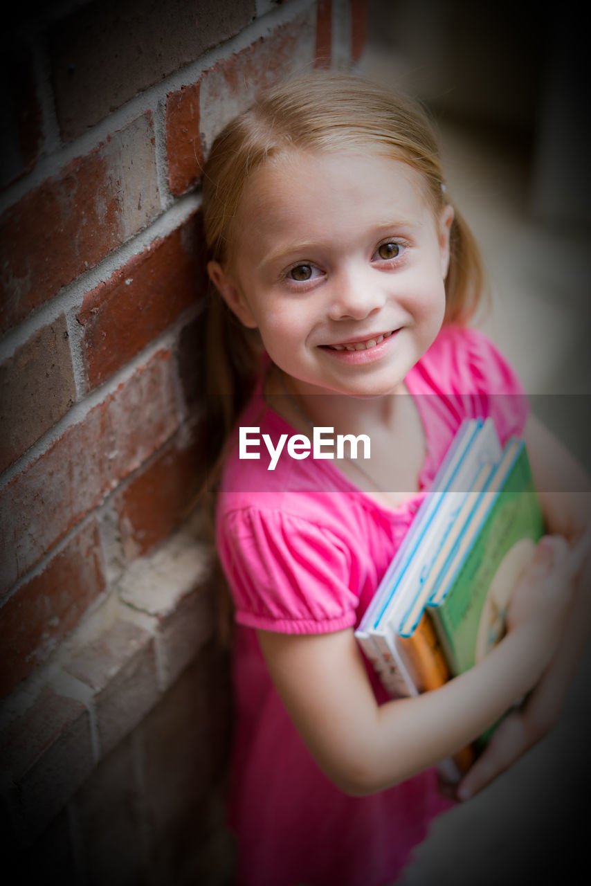 Portrait of smiling girl holding books by brick wall