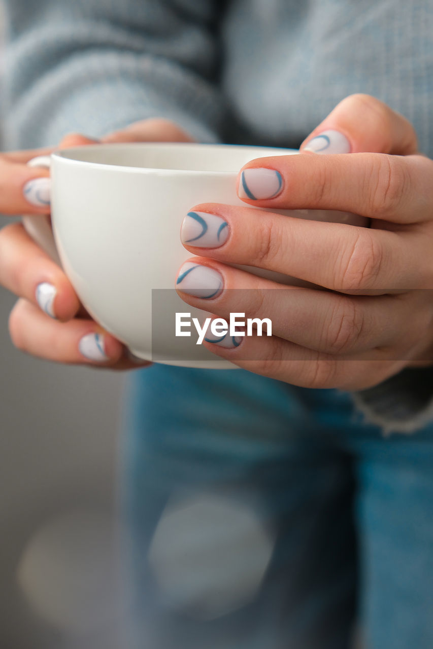 Manicured female hands holding white coffee cup with stylish blue nails and minimalistic design. 