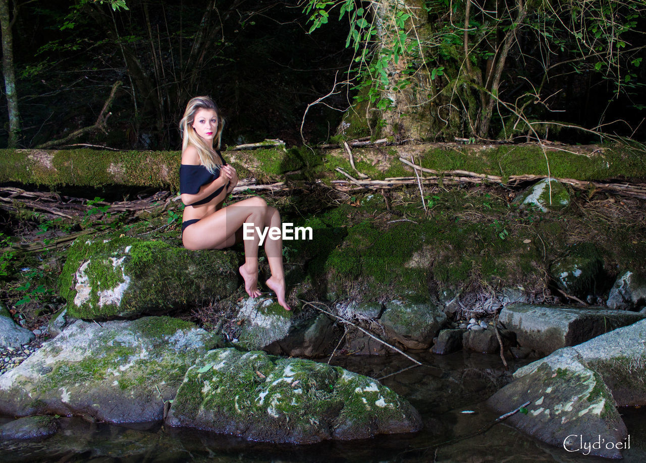 Full length of beautiful woman sitting on rock at lakeshore in forest