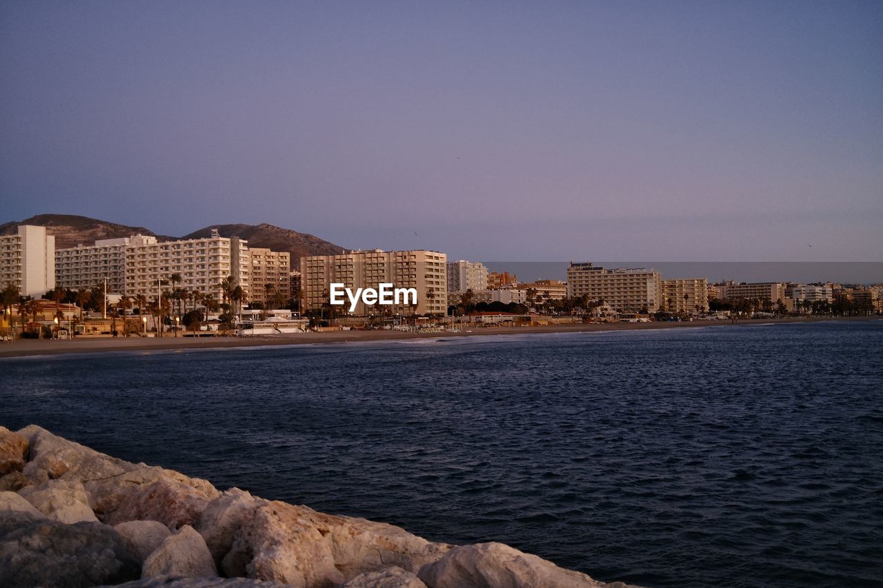 architecture, horizon, building exterior, city, built structure, water, dusk, sky, coast, building, sea, evening, sunset, nature, beach, cityscape, travel destinations, shore, land, ocean, skyline, landscape, bay, urban skyline, travel, no people, reflection, clear sky, outdoors, night, residential district, office building exterior, city life, body of water, coastline