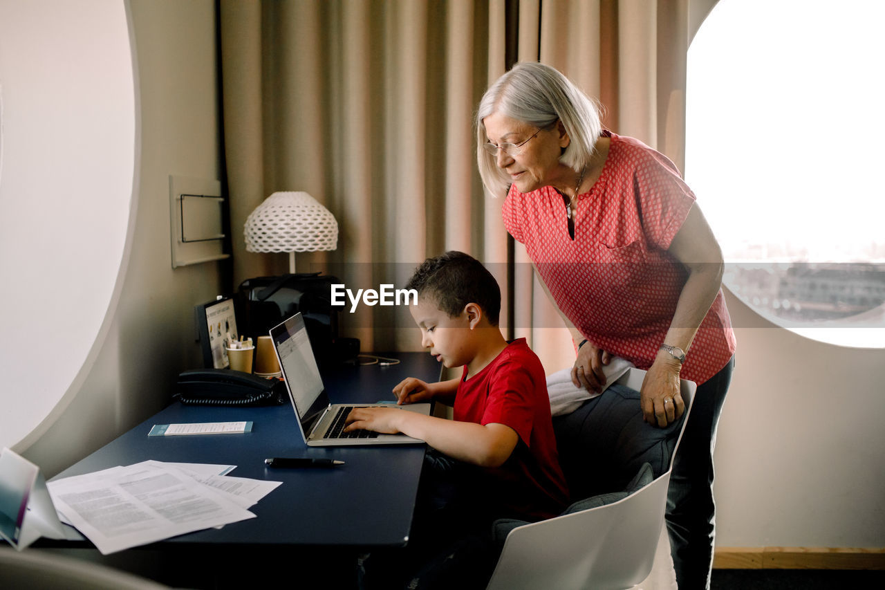 Grandmother looking at grandson using laptop while sitting by table in hotel room
