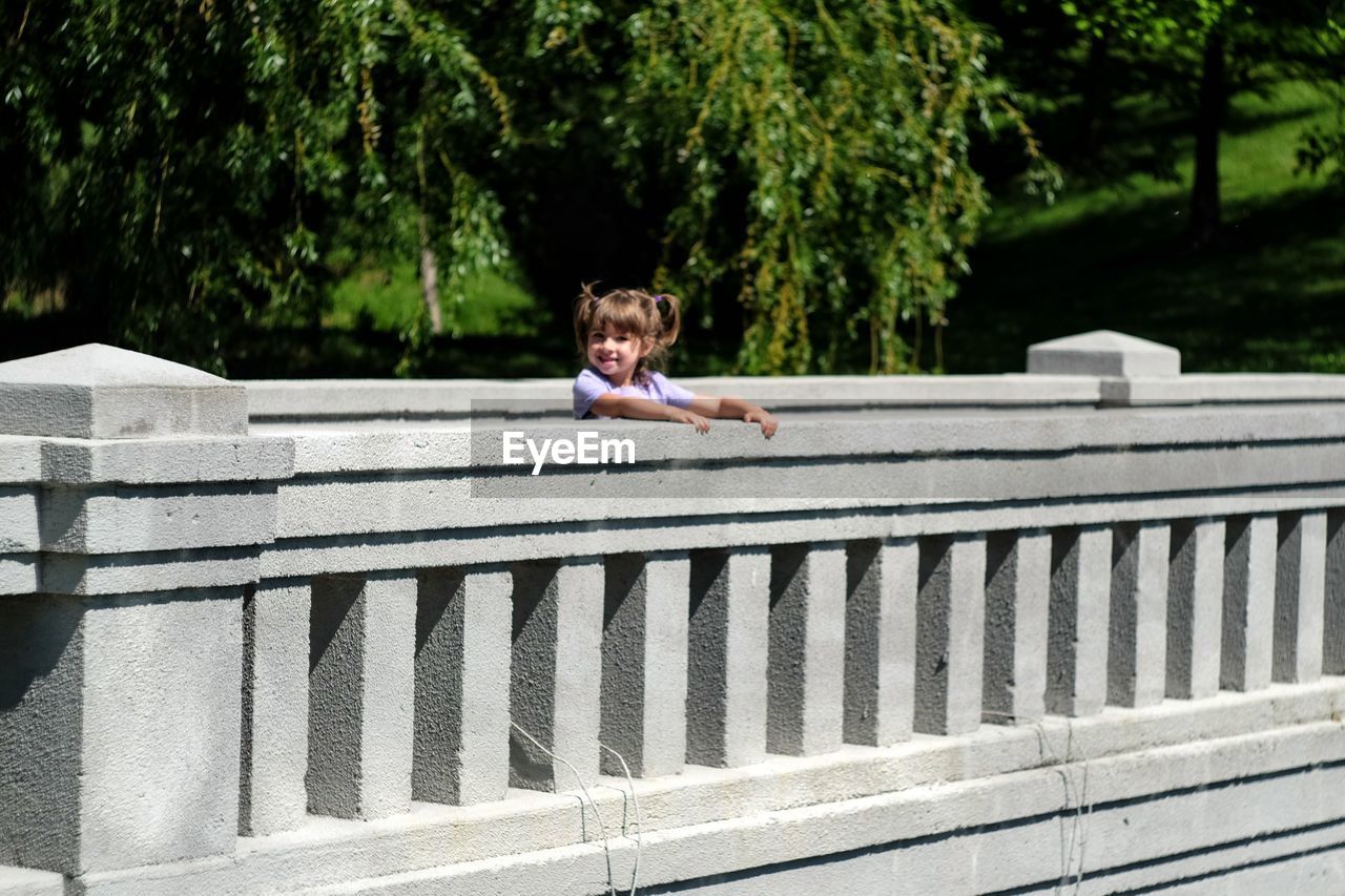 Smiling girl standing by railing while looking away on sunny day