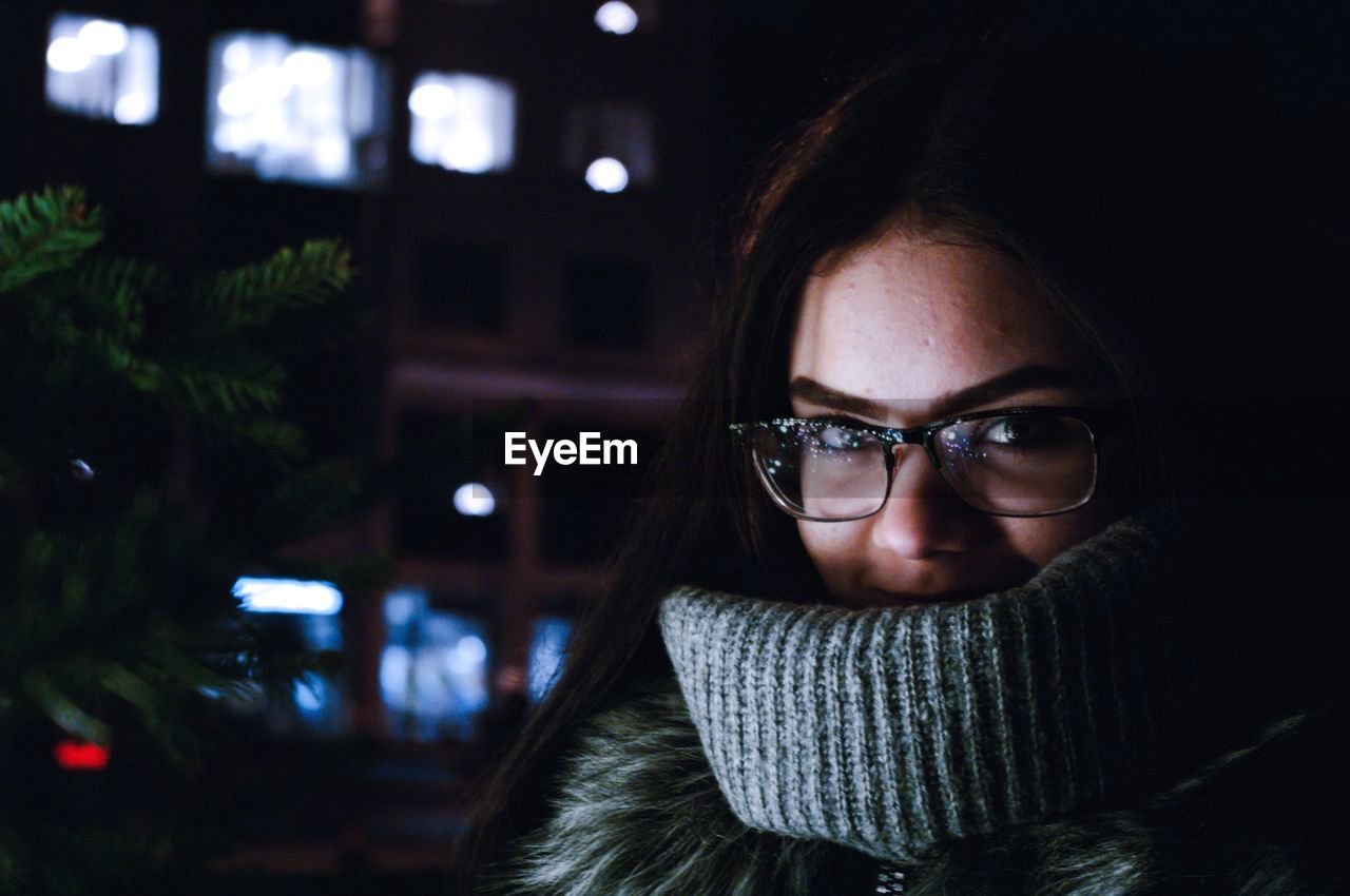 Close-up portrait of young woman in warm clothing standing against building at night