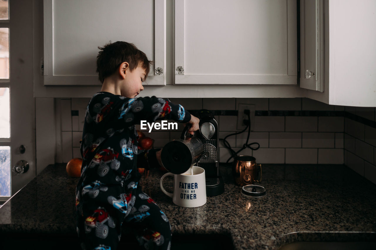 Young boy sitting on kitchen counter in pjs making coffee for dad