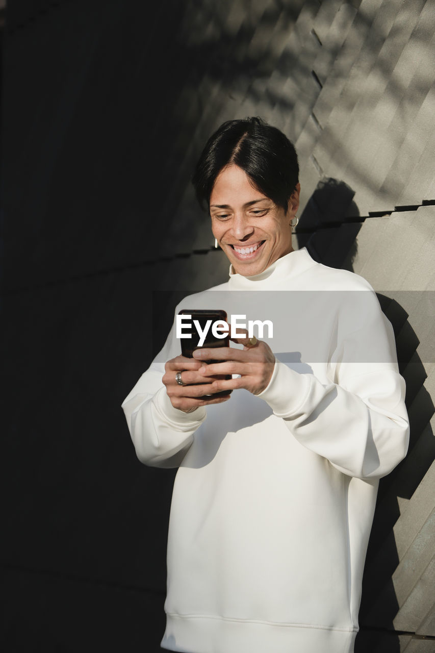 Smiling asian man in white jumper standing on street concrete wall and reading messages on smartphone