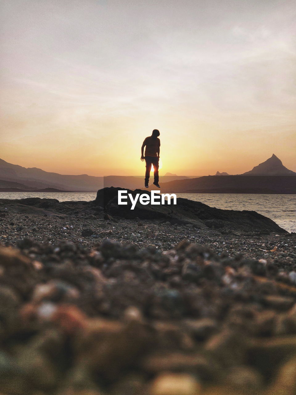 MAN STANDING ON ROCK AT BEACH DURING SUNSET