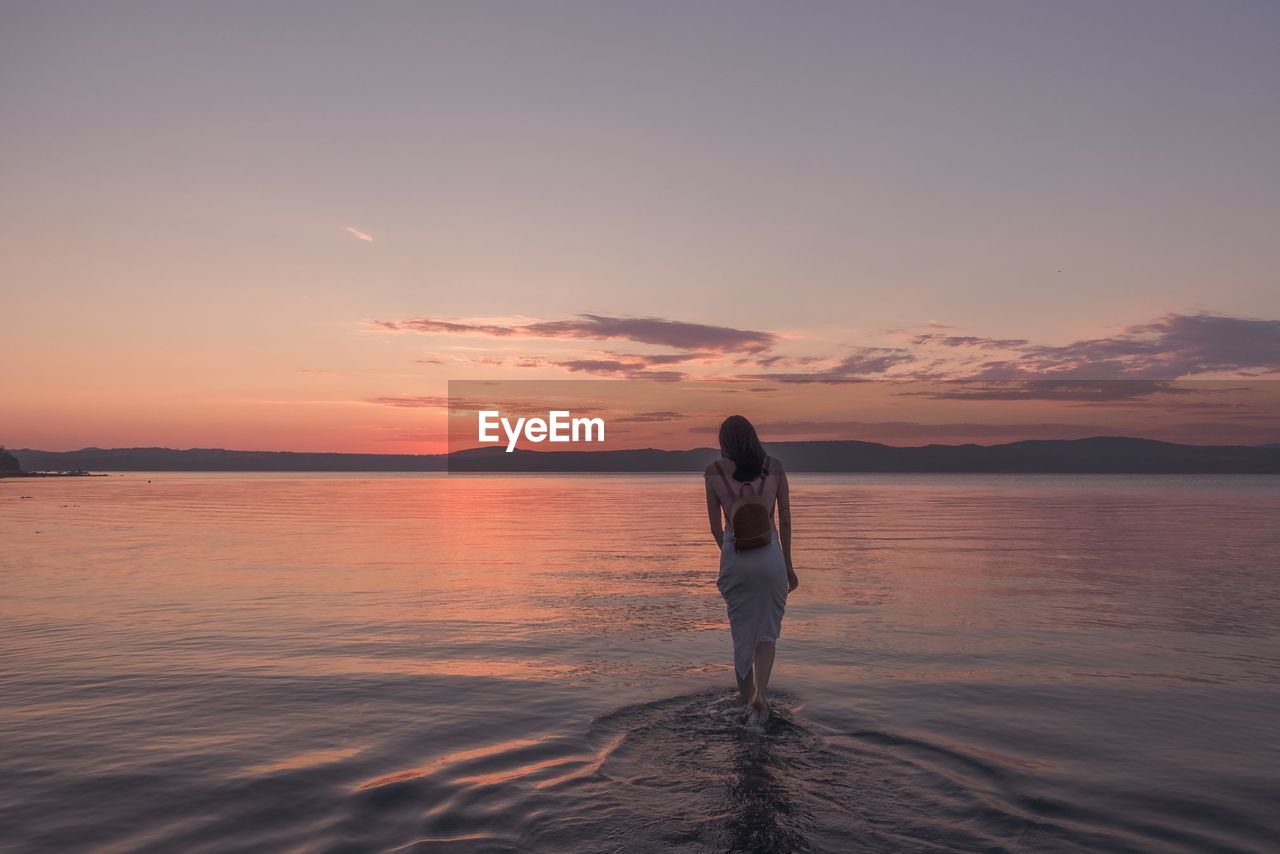 Rear view of woman walking in lake against sky during sunset