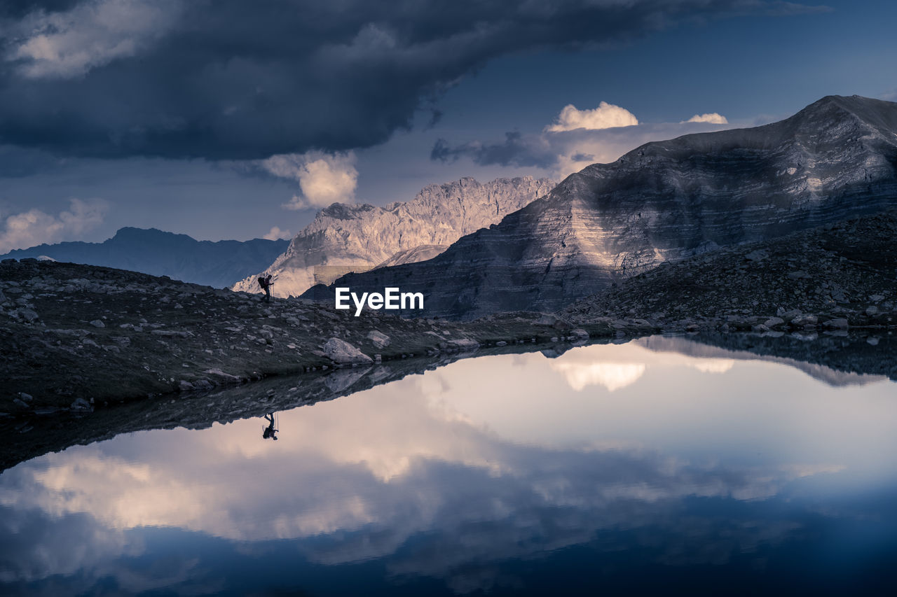 Alpine lake with cloudy sky and trekker reflecting on watee
