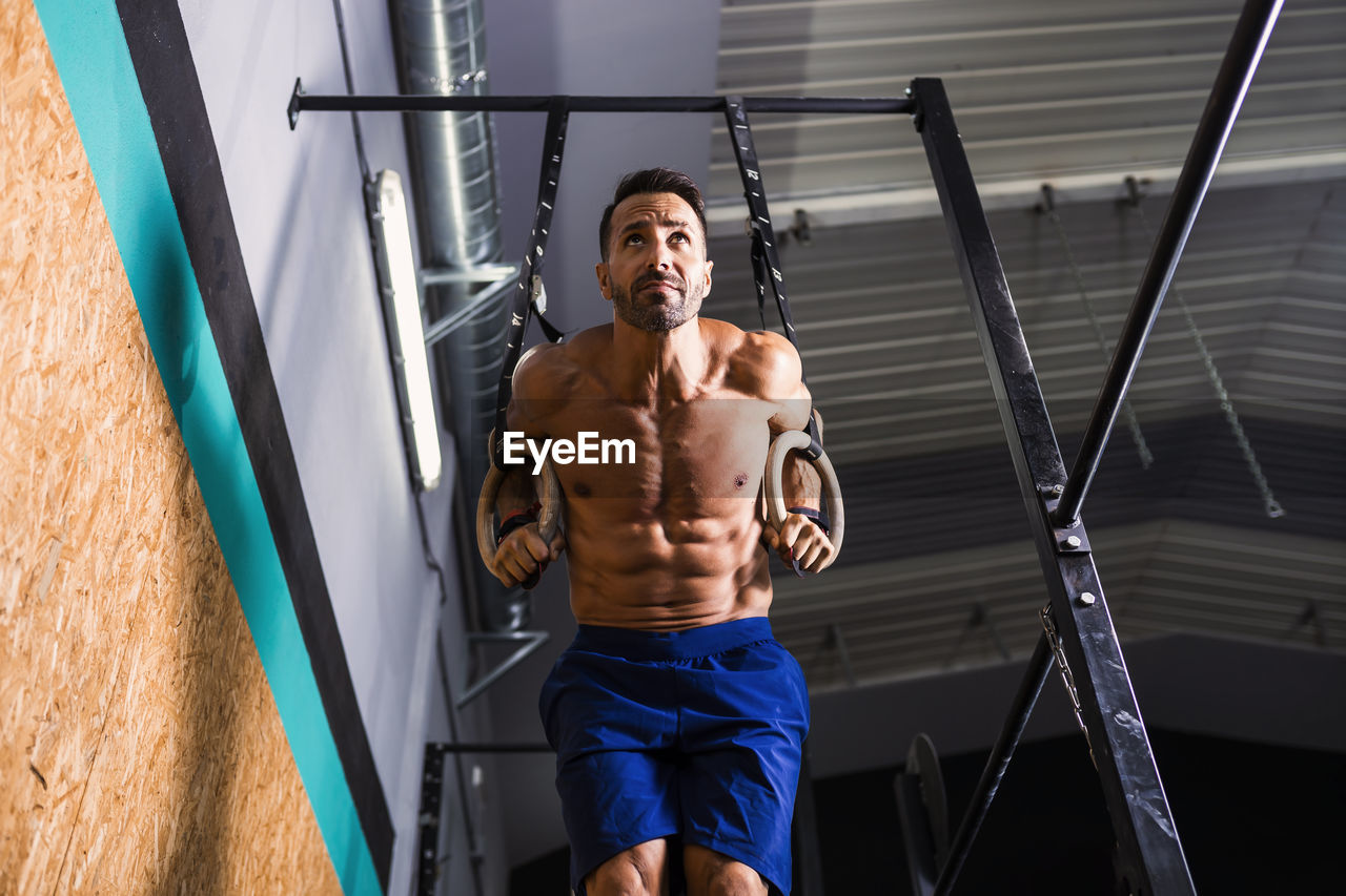 Shirtless male athlete exercising on gymnastic rings in gym