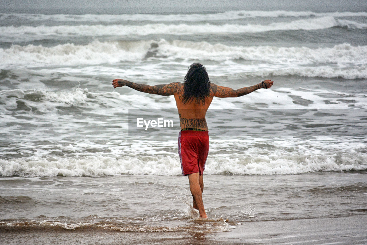 Rear view of shirtless man with arms outstretched wading in sea