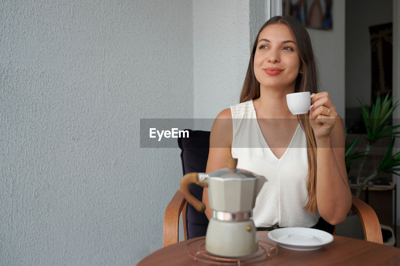 Beautiful young woman enjoying drinking italian coffee espresso sitting on terrace at home. 