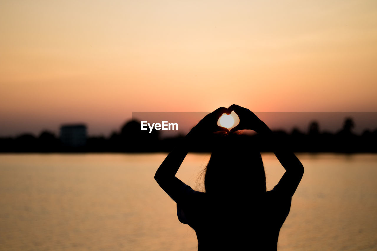 Silhouette woman making heart shape with hands by lake against sky during sunset