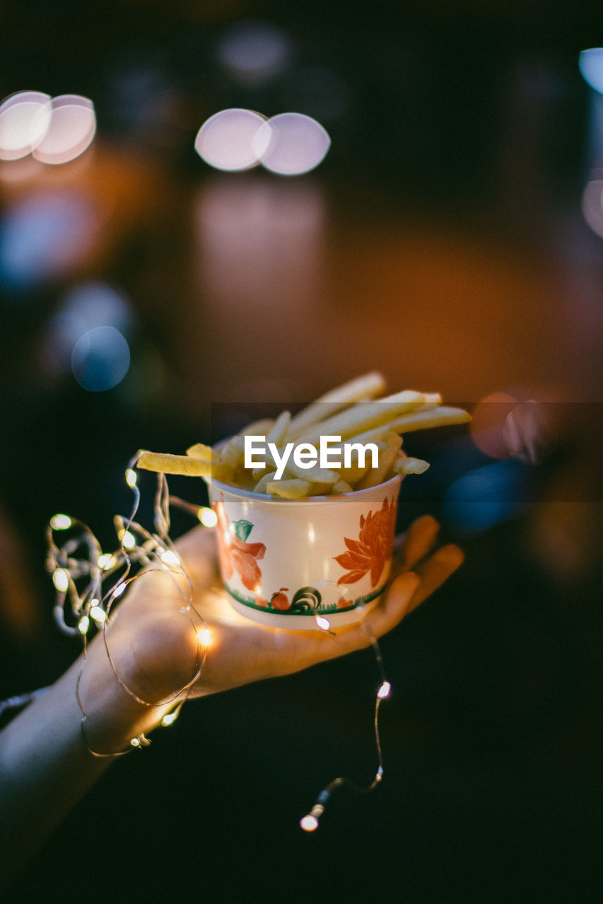 Cropped image of hand holding french fries in bowl with lighting equipment during night outdoors