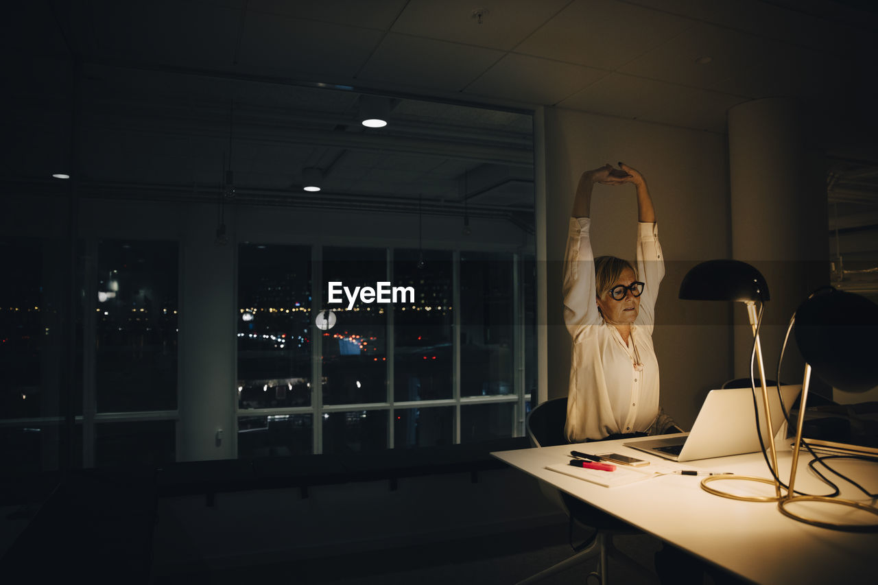 Exhausted businesswoman stretching with arms raised while working late in illuminated coworking office