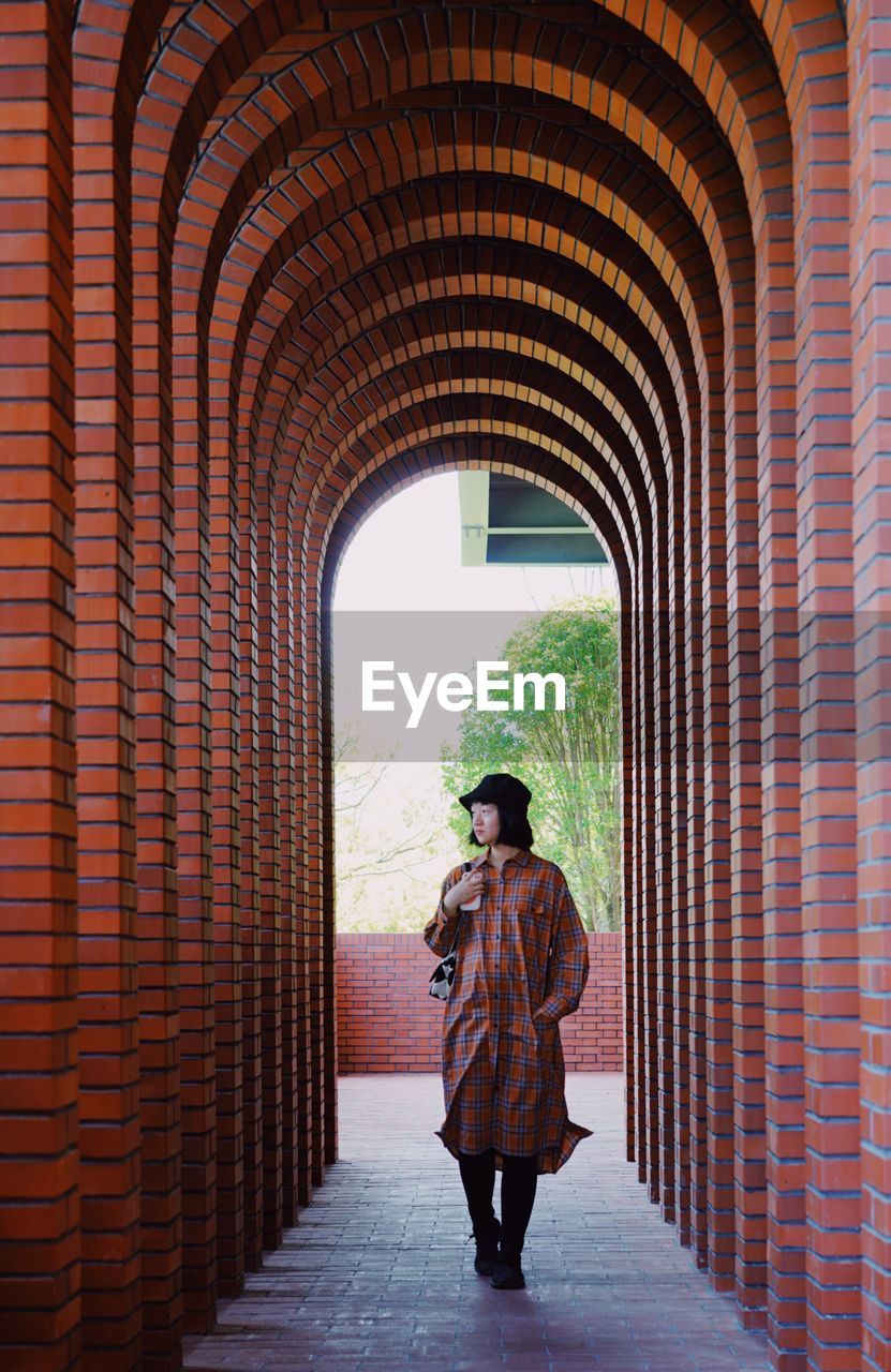arch, one person, full length, architecture, adult, brick, standing, built structure, brick wall, day, temple, clothing, history, the way forward, the past, wall, young adult, walking, arcade, casual clothing, men, indoors, travel destinations, red, lifestyles, leisure activity, travel, corridor, rear view, person, tourism, looking