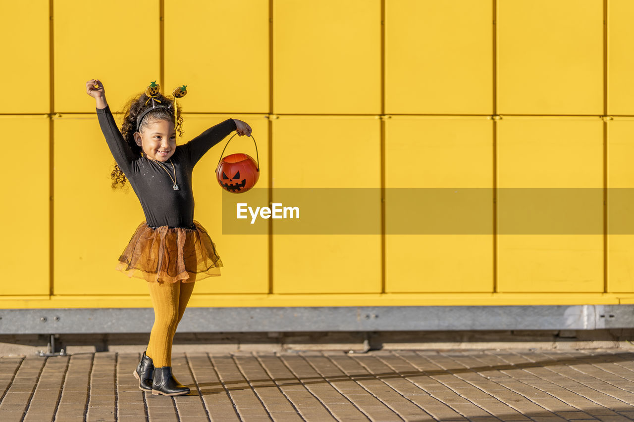 Smiling girl dancing with halloween flower pot and headband against yellow checked pattern wall