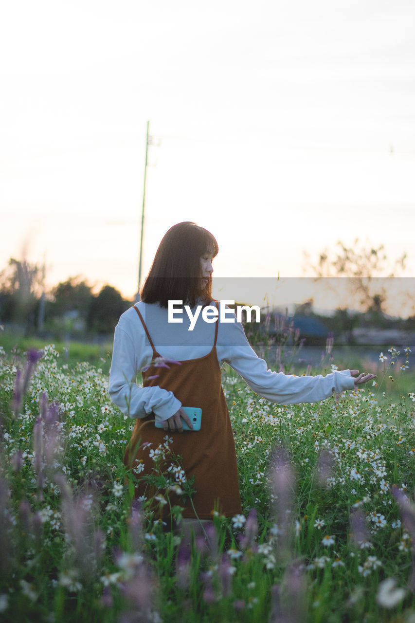 REAR VIEW OF WOMAN STANDING ON FIELD BY FLOWERING PLANTS