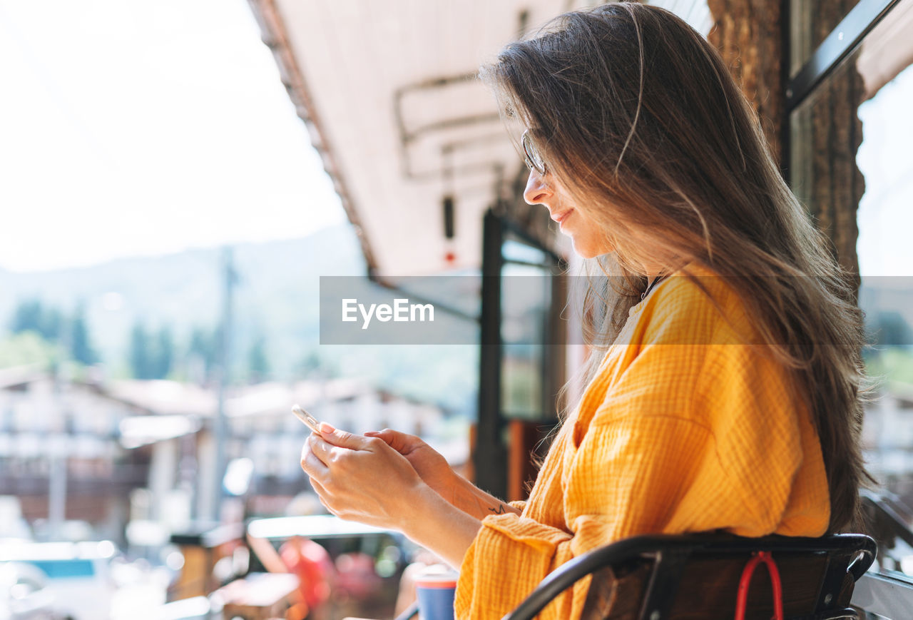 Young woman with long hair sunglasses and yellow longsleeve with mobile phone in hands in cafe