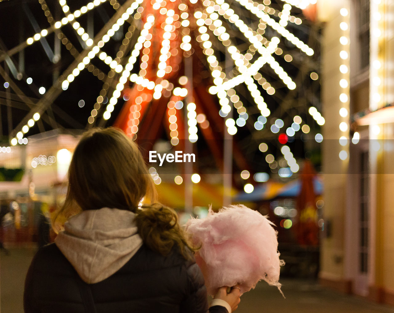 Rear view of woman holding cotton candy against illuminated ferris wheel at night