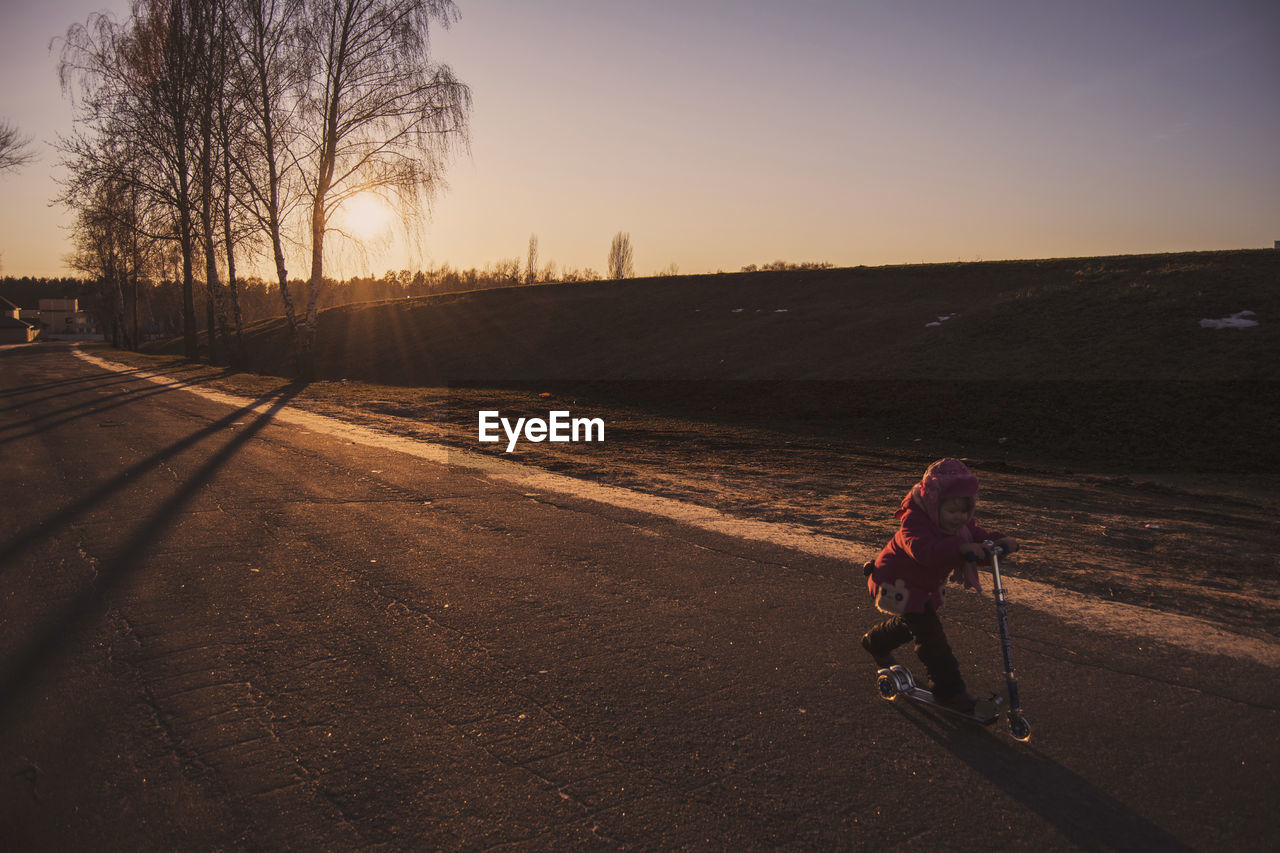 Girl riding push scooter on road against clear sky during sunset